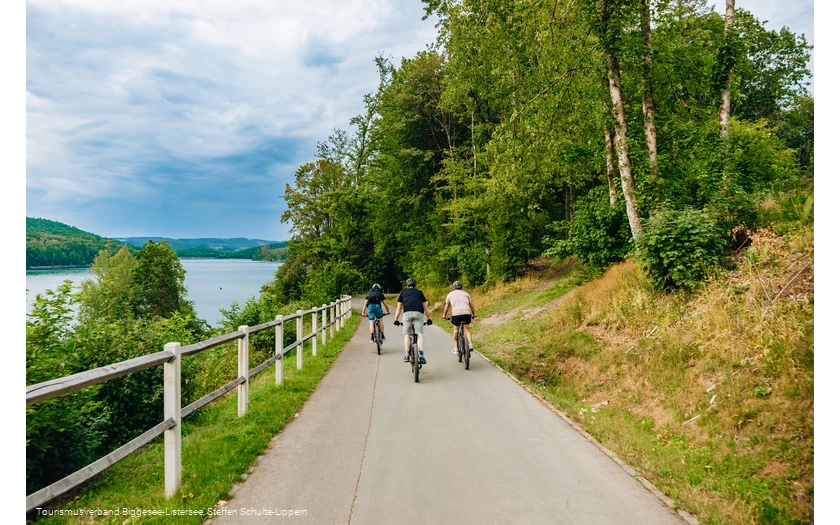 Drei Männer fahren entlang des Bigge-/Listersees auf ihren Fahrrädern weg von der Kamera. Der Himmel im Hintegrund ist wolkenverhangen.