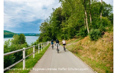 Drei Männer fahren entlang des Bigge-/Listersees auf ihren Fahrrädern weg von der Kamera. Der Himmel im Hintegrund ist wolkenverhangen.
