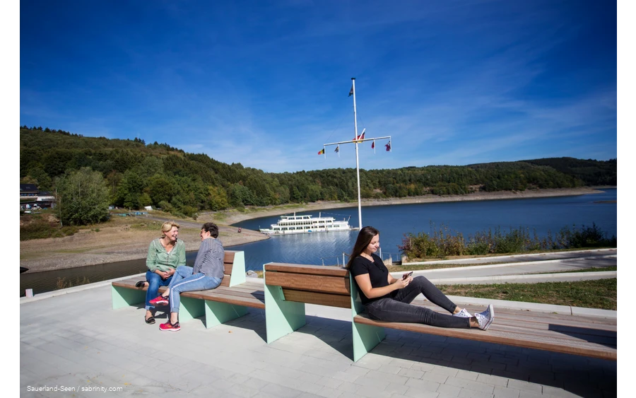 Im Vordergrund sitzen drei Frauen auf Bänken, zwei unterhalten sich und eine ist am Handy. Im Hintergrund ist der Bigeesee mit dem Schiff.