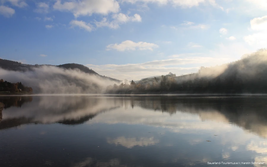 Diemelsee im November mit Nebel über dem Wasser und blauem Himmel, der sich im Wasser spiegelt.