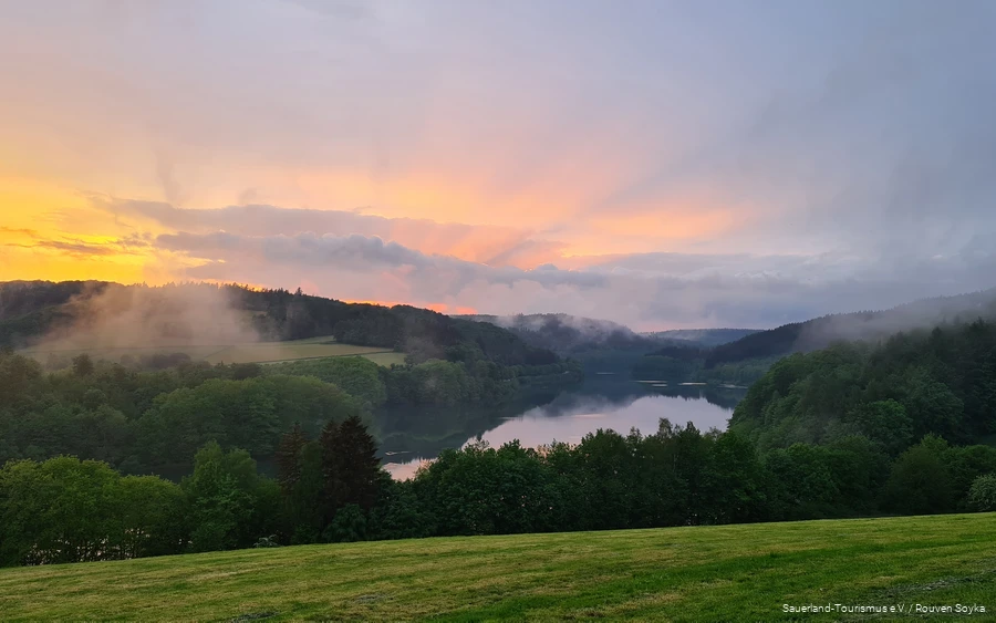 Über dem Hennesee und den Bäumen steigt Nebel auf und der Himmel kreiert mit Wolken und der unterhehenden Sonne ein Farbspektakel.